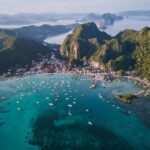 Panoramic aerial drone shot of numerous tour boats anchored in a sweeping turquoise bay surrounded by dense green coastal mountains in El Nido, Palawan.