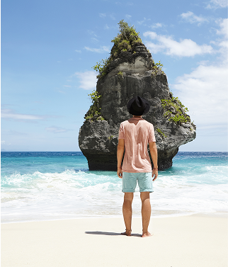 A traveler standing on a pristine white sand beach looking out at a unique, towering limestone rock formation in the turquoise sea of El Nido.