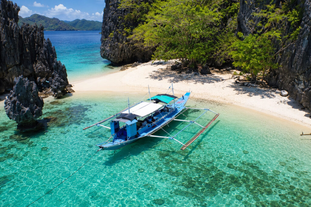 Um barco de relevo tradicional filipino azul e branco ancorado em uma praia de areia branca ao lado de águas turquesa e penhascos de calcário escuro.